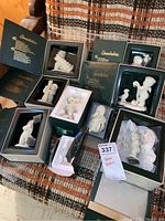 Photo showing eight Department 56 Snowbabies figurines in original boxes on a patterned brown and cream fabric surface.