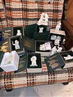 Display of multiple Snow Babies figurines on a patterned chair, several still in their original dark green boxes with gold text.