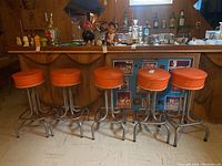 Five vintage orange vinyl covered metal barstools arranged in front of a wooden bar with various items on top.