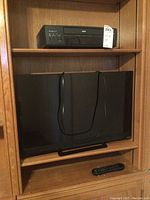 Photo showing the Vizio television with remote control, placed on a shelf inside a wooden cabinet.