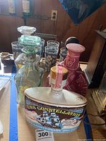 Photo shows a group of whiskey decanters including the USS Constellation 175th anniversary decanter in front, vintage glass set with metal holder and seven floral pattern glasses behind them.
