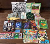 Full assortment of MaxSold unloved items displayed on floor including scout clothing, comic books, art books, DVDs, cards, and collectibles.