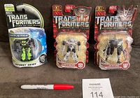 Front view of three unopened Transformers action figures on cards showing Autobot Skids, Sideswipe, and Jetfire in packaging with maxsold lot number and red marker for scale.