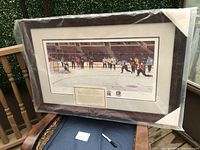 Photo showing the framed print in original plastic with cardboard corner protectors, featuring a hockey practice scene on ice with players in jerseys.