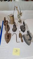 Photo of antique sad irons, metal fuel pot, meat grinder, cast iron shoe form, and other tools displayed on a table.