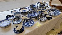 Wide angle showing full collection of Blue Willow transferware including multiple plates, cups, bowls, and saucers laid out on a table.