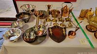 Overview of the collection on a display table including silverplate bowls, brass trophies, candelabras, copper kettle and measuring cup, and a wooden plaque.