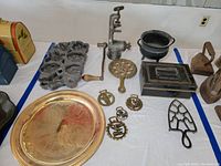 Overview of the lot showcasing the gold plated platter, cast iron flower baking mold, manual meat grinder, small cast iron pot, vintage tin box, cast iron trivets with animal motifs, antique irons, and a cast iron trivet