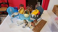 Overview photo showing various colored glass bottles, musical wind-up egg, metal doll chairs, Delft miniatures, polished stones, fossils, shells and a cribbage board arranged on a table.