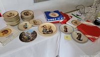 Photo showing multiple stacks of Hummel collector plates on a table with some boxes and packaging nearby.