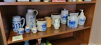 Shelf display of 11 artisan pottery pieces including jugs, crocks, steins, and small vessels with salt glaze and cobalt blue blueberry basket decoration.