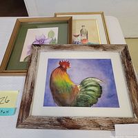 Photo showing three framed watercolor paintings by Elaine Stedman featuring a rooster, flowers, and a still life with wine and fruit.