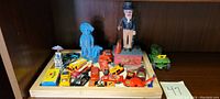 Photo showing multiple toy cars, Uncle Sam bank figurine, cast iron motorcyclist, and John Deere tractor on a wooden tray.