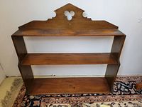 Full front view of the vintage dark finish wood wall shelf standing on a patterned rug against a white wall, showing shelves and decorative top.