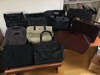 Full view of the lot showing satchels, backpacks, rolling case, and lap desk on a wooden table against a white wall.