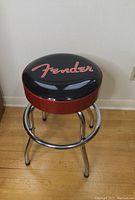 Front angled view of the Fender guitar stool showing the black vinyl seat with red Fender logo, red sparkle trim wrapping, and chrome legs with footrest.