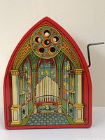 Front view of music box showing arched red top and decorative church organ lithograph with stained glass windows and central organ pipes.
