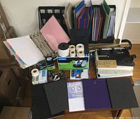 Photo of all items in lot arranged on table showing binders, file folders, label maker and tapes, hole punch, stapler, tape dispenser, calculator, and papers