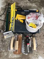 Mastercraft wet saw, manual tile cutter with instructional card, bucket of tile spacers, and tile trowels on concrete floor
