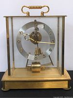 Front view of the vintage mantel clock showing clear glass panels, gold-tone frame, circular visible clock dial with gold Arabic numerals and hands, pendulum in center.