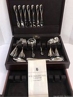 Full view of the open case showing the entire stainless steel flatware set inside including knives secured in the top section and various forks, spoons, salad utensils, and serving spoons in the bottom section. A WMF pamphlet is also displayed inside the case.