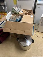 View showing a cardboard box containing 12 Ball half-pint mason jars packed in original packaging, sitting on top of a metal pressure cooker with lid and handles.