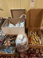 Wide view showing boxes containing china cups and saucers wrapped in bubble wrap, Bakelite flatware with assorted handles, and colored glass goblets.