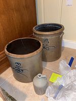 Two stoneware crocks side by side showing relative size and visible crack in smaller crock, with small ceramic jug in front.
