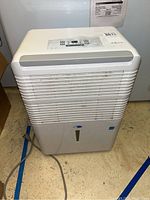 Full front view of white Whynter dehumidifier on basement floor, showing air intake grill and power cord.