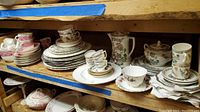 Wide view of the shelf showing multiple stacks of plates in various sizes, cups stacked on saucers, a tall floral teapot, and cream and sugar containers.