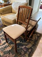 Three wood folding chairs stacked with beige upholstered seats and stained wooden frames, shown in living room space on patterned rug.
