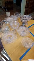 Photo of ten or more clear EAPG glass items arranged on a wooden table including pitcher, candy dishes, lamp base, cake plates, and fruit bowls.