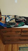 Photo of a stack of folded quilts and quilt squares on a wooden dresser, showing a variety of colors and patterns including plaids and patchwork.
