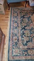 Photo showing part of the wool area rug with floral and leafy patterns and bordered design on wooden floor near furniture.