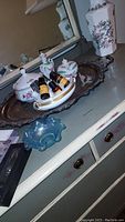 Wide view of the silver plated tray holding porcelain bowls, perfume bottle, small amber bottles, blue glass bowl, and bird figurine on a dresser.
