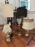 Four table lamps on hardwood floor in room with wood furniture and window, showing ceramic and metal bases with various shades including glass and fabric.