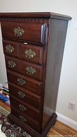 Full view of the tall wooden vanity dresser with eight drawers and brass handles against a wall.