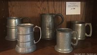 Five beer steins on shelf; three appear hand-hammered pewter, two smooth metal. One pewter stein has music box in base.