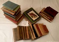 Three stacks of books shown from above on a flat surface. Books are worn, with some having cloth or hard covers, others paperbacks with visible texture and color fading.