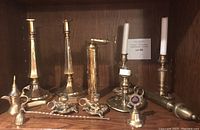Full assortment of brass items including three sets of candlesticks, candle snuffer, spice grinder, and bell displayed on wood shelf.