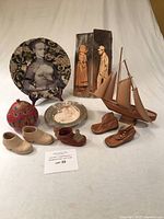 Full lot image showing model sailboat, decorative Esther Baker plate, red lidded dish, various children's shoes including leather and ceramic, knitted booties, stainless steel child's dish, and wooden carved figures