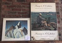 Framed poster signed by A.Y. Jackson with significant water damage and framed Group of Seven print side by side against brick wall.