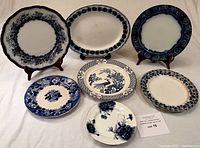Seven assorted blue and white porcelain and semi-porcelain plates displayed standing and lying down showing various floral and patterned blue borders and centers.