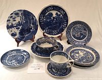 Full view of seven blue and white ceramic pieces including plates on stands and cups with saucers arranged on a white surface, showing detailed patterns and designs