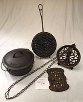 Overall grouping showing Dutch oven, perforated pan, decorative and rectangular trivets, twisted metal tongs and lifting tool