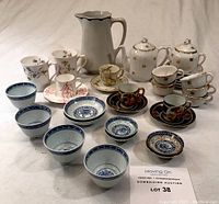 Lot of porcelain items with teapot, sugar dish, cups, saucers, dipping bowls and water jug displayed on a white surface.
