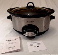 7 quart Crock-Pot slow cooker with stainless steel exterior, control dial, and accompanying owner's guide and lot card visible.