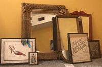 One large gilt gold framed mirror with detailed ornate edges and one medium gilt gold framed mirror with simpler edges. One red painted wood framed mirror with decorative top edge. Four assorted framed art prints including a red shoe fashion print and vintage style prints, plus glass wind chimes with blue and green glass droplets.