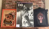 Three framed artworks placed side by side on a brick hearth; left is painting on burlap, center is Charlie Parker poster, right is portrait of young woman.