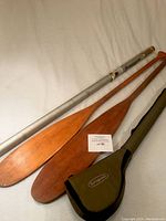 Two wooden canoe paddles and two fishing rod cases laid out on a white background, showing paddles with natural wood finish and cases including one aluminum tube and one fabric Springbrook case.